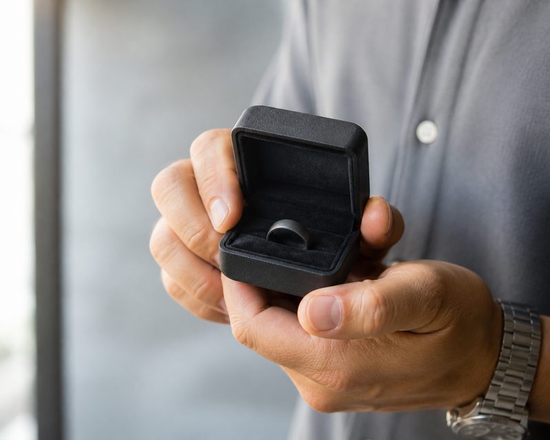 Man's hands holding an open ring box with a dark wedding band inside