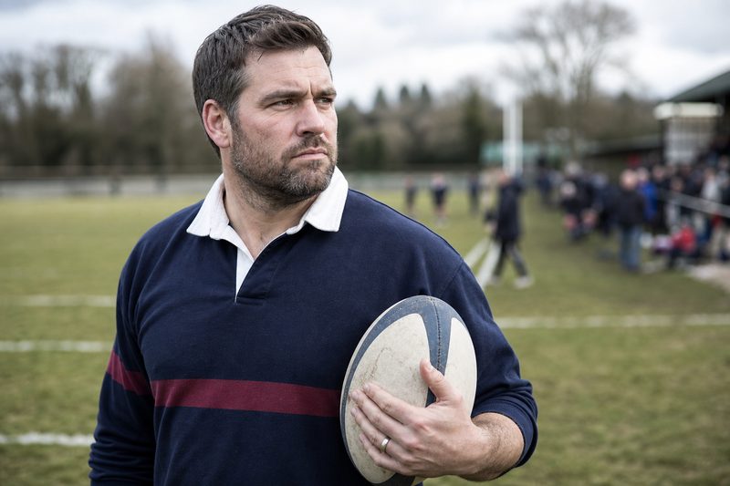 Man in a rugby jersey on the sideline holding a rugby ball, wedding band visible
