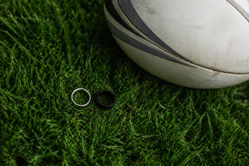 Metal wedding band and black silicone ring beside a rugby ball on grass
